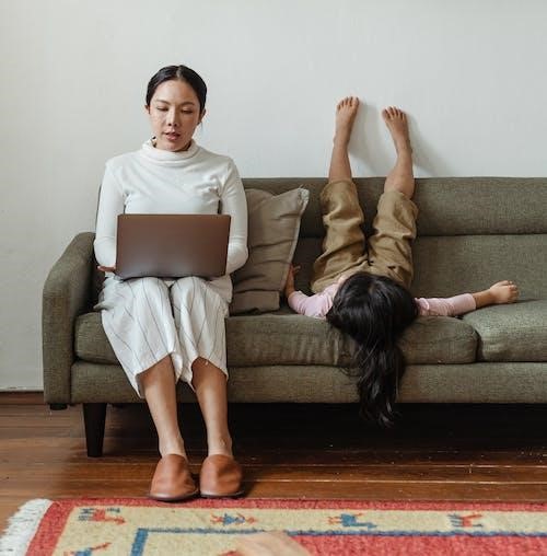 Parent using laptop and child laying upside down on sofa